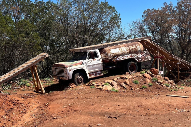 An old, rusty dump truck is positioned on a dirt mound surrounded by trees, with a wooden ramp leading up from the ground beside it. The truck has paint peeling off and a partially raised bed. The area appears natural, with patches of grass and scattered rocks. Bright blue skies are visible in the background.