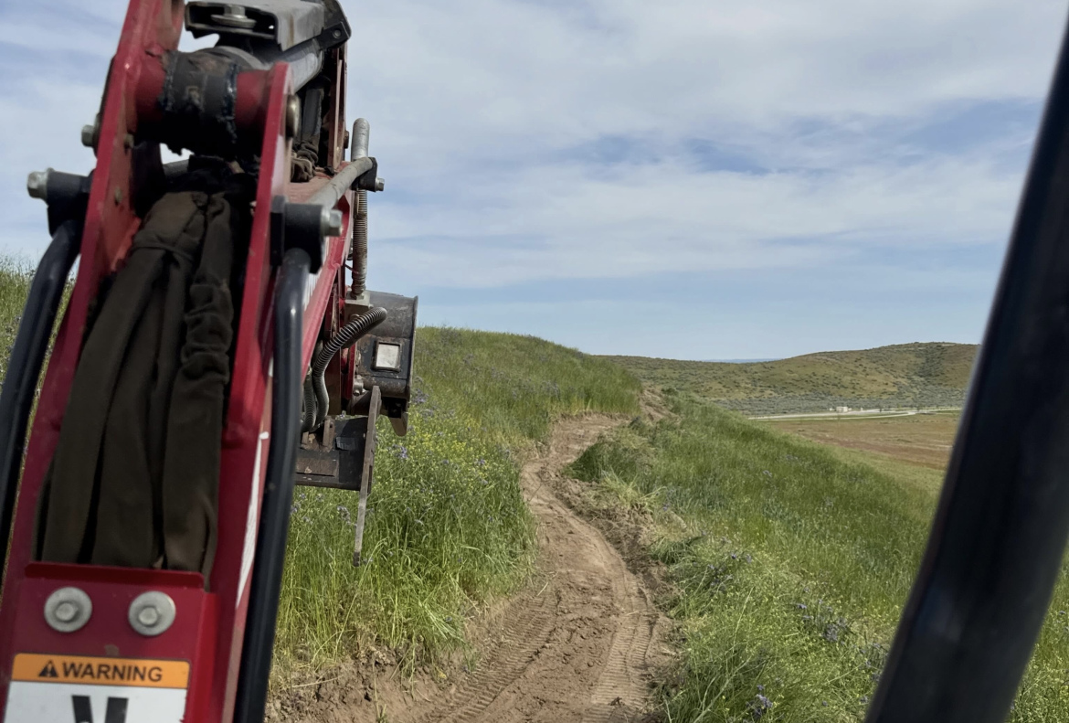 A close-up view of a digging machine's arm positioned over a dirt path lined with green grass and wildflowers, leading toward rolling hills under a partly cloudy sky.