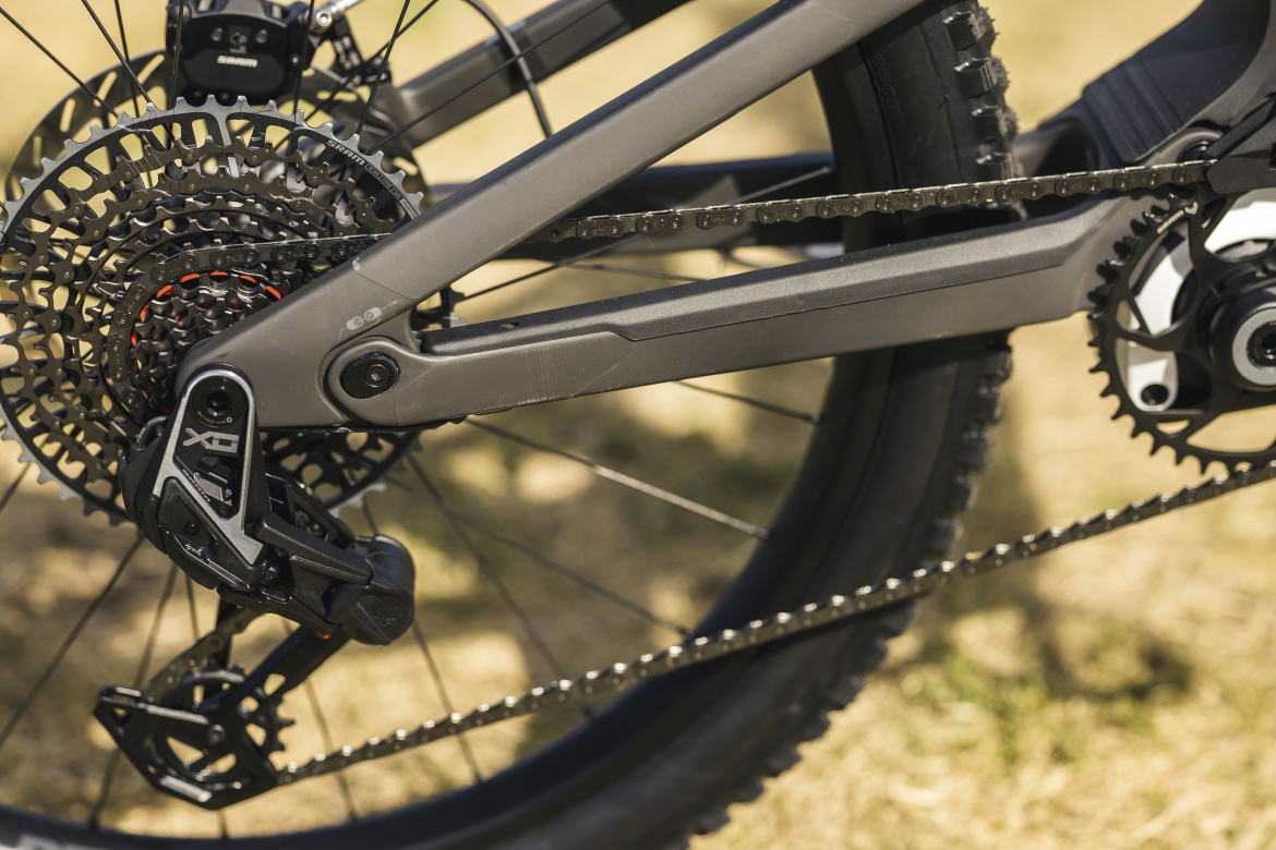 Close-up view of a mountain bike's drivetrain, showcasing the rear derailleur, chain, and gears against a blurred natural background. The components are primarily black with metallic accents, highlighting the bike's design and mechanical features.