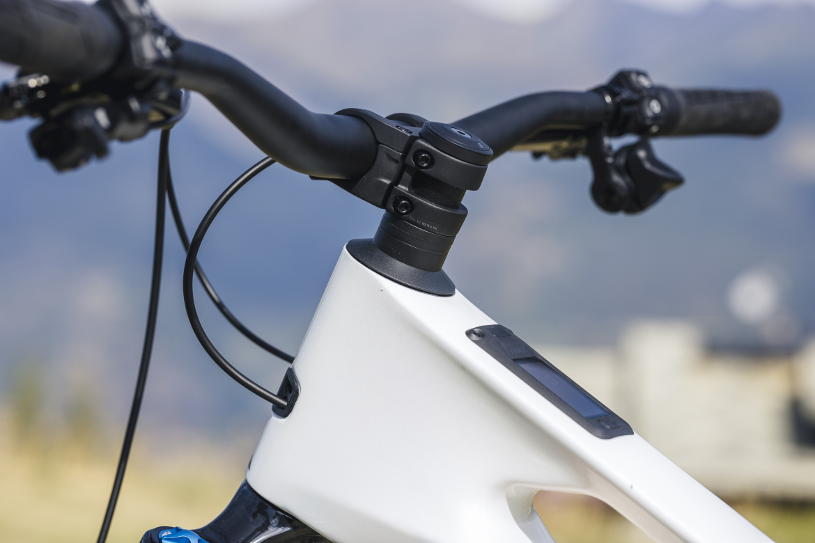 Close-up view of a mountain bike's handlebar and headset with a sleek white frame. The handlebars are black and equipped with controls, while the bike features an integrated display on the top tube. The background shows blurred mountain scenery, emphasizing the outdoor cycling environment.