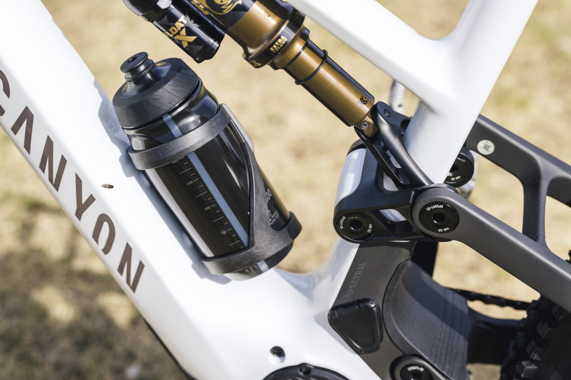 Close-up of a mountain bike frame featuring a black water bottle mounted beside the rear shock. The bike has a sleek, white design, with visible branding and mechanical components. The background is blurred grass, suggesting an outdoor setting.