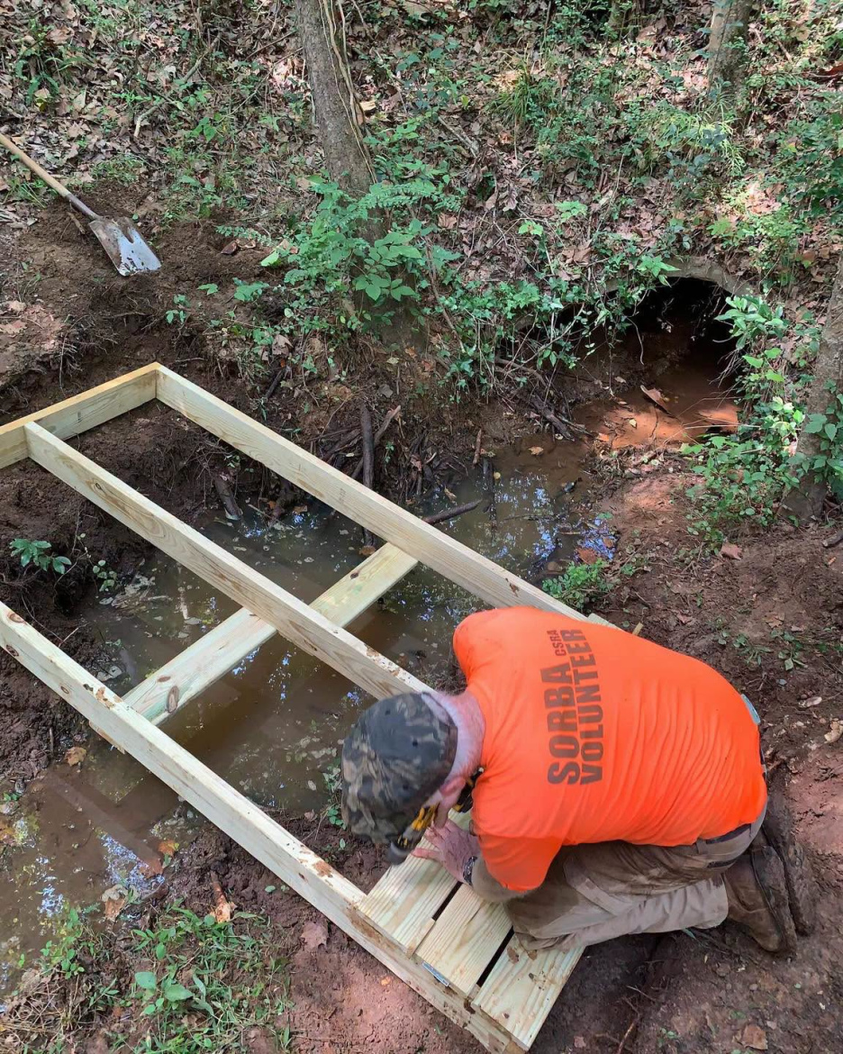 A person in an orange shirt is kneeling next to a partially built wooden structure over a small stream in a wooded area. The surrounding environment features green vegetation, soil, and a drainage tunnel visible in the background. A shovel is placed nearby on the ground.