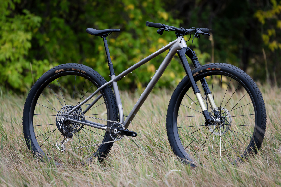 A mountain bike is positioned on grassy terrain, featuring a sleek metallic frame, wide tires, and a front suspension fork. The background showcases blurred greenery, suggesting an outdoor environment.