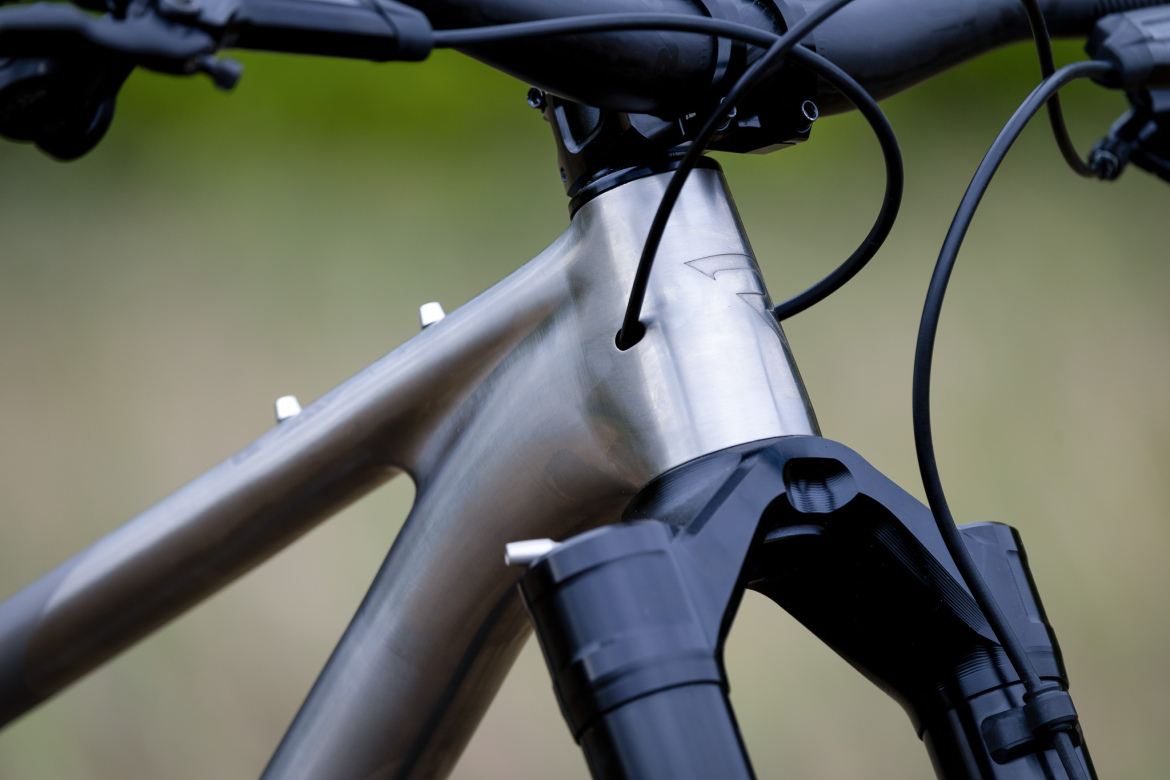 Close-up of a mountain bike frame showing the silver metal head tube and black fork assembly. The image highlights the detailed design and construction of the bike's components, with visible cables and a blurred natural background.