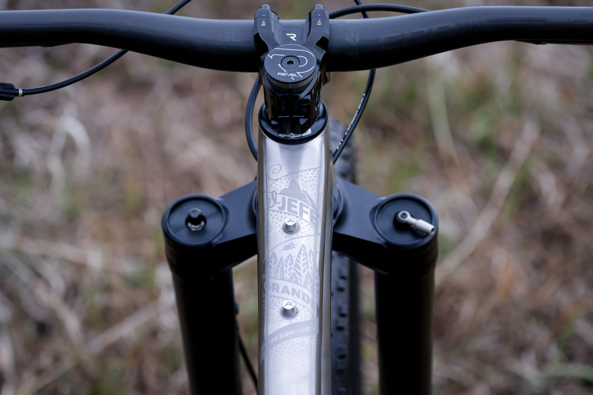 Close-up view of a mountain bike's handlebars and frame, featuring a metal top tube with intricate designs and text. The bike's stem displays a logo, and there are two black adjuster knobs on the front. The background includes blurred, earthy tones of grass and foliage.