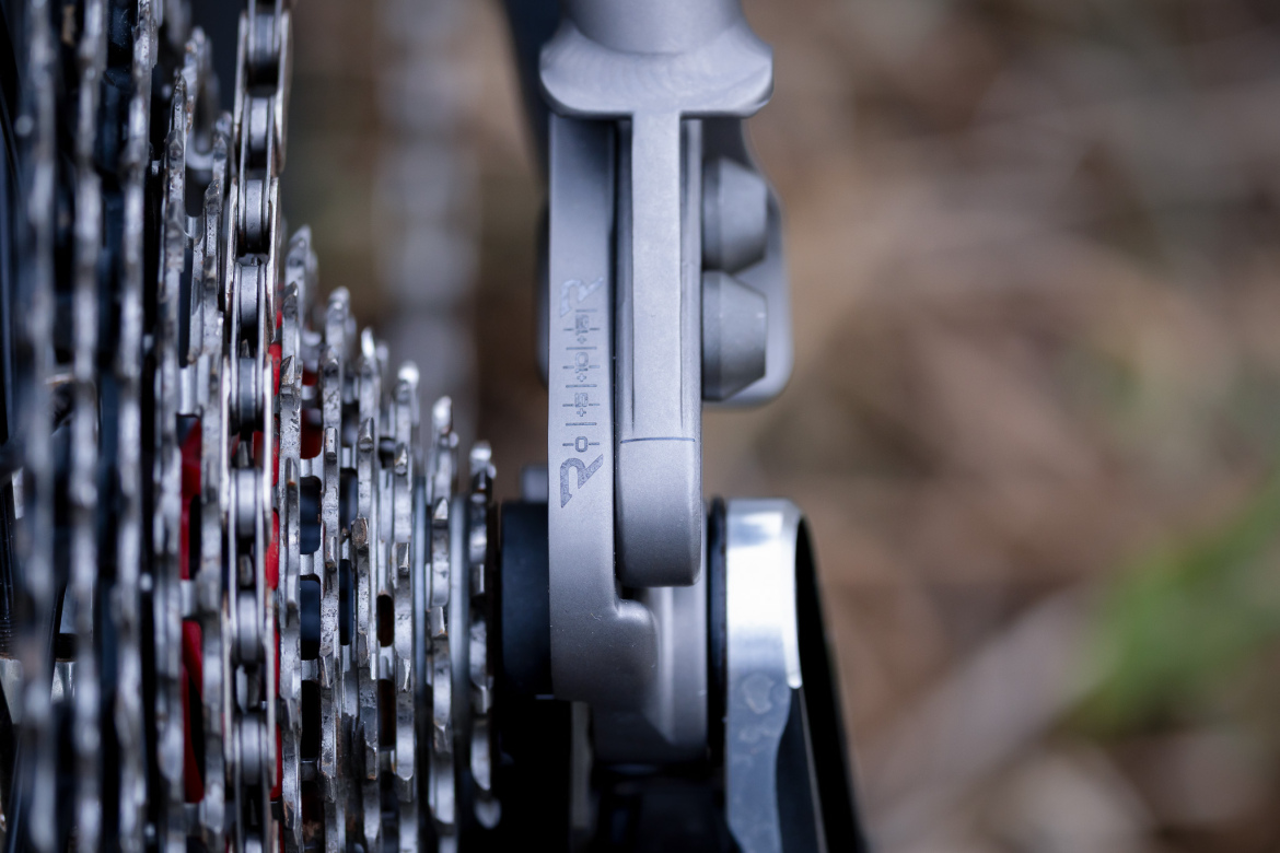 Close-up of a bicycle drivetrain, featuring a cassette with metallic gears and a gray derailleur. The image shows detailed textures and the precision engineering of the components, with a blurred background of natural surroundings.