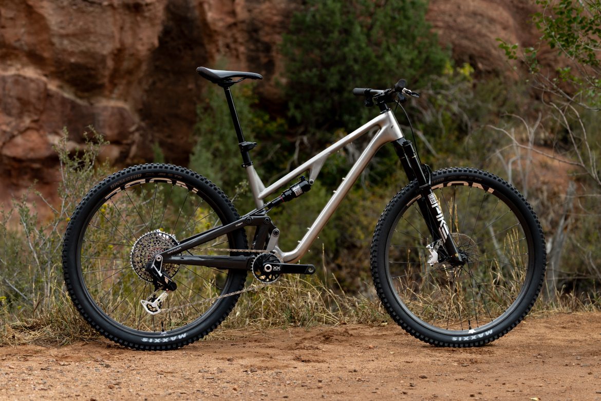 A modern mountain bike with a silver frame sits on a dirt path surrounded by greenery and rocky terrain. The bike features thick tires and a suspension system, positioned upright for a clear view of its design and components.