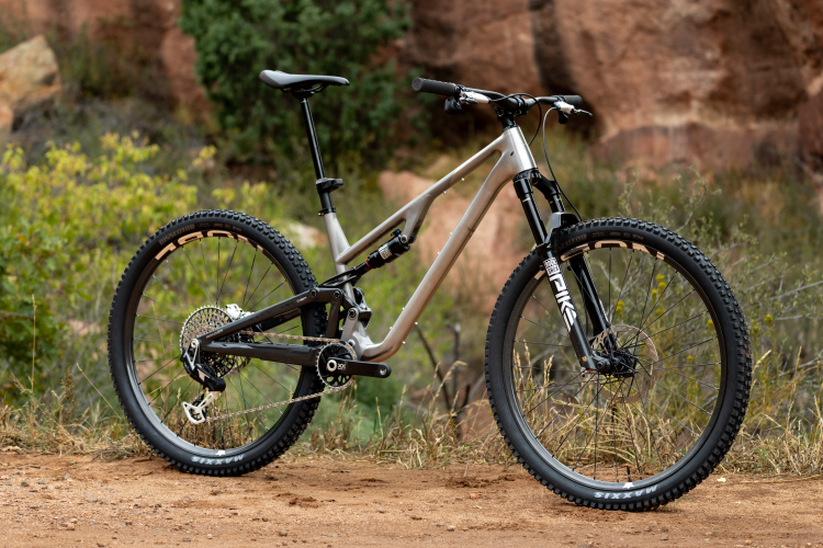 A sleek mountain bike with a silver and black frame, equipped with a front suspension and thick, knobby tires, positioned on a dirt path surrounded by greenery and rocky terrain.