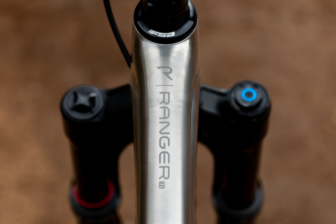 Close-up view of a mountain bike frame featuring the brand name "Ranger" prominently displayed. The bike's suspension components are visible, with one labeled FSA, set against a blurred brown background, suggesting an outdoor setting.
