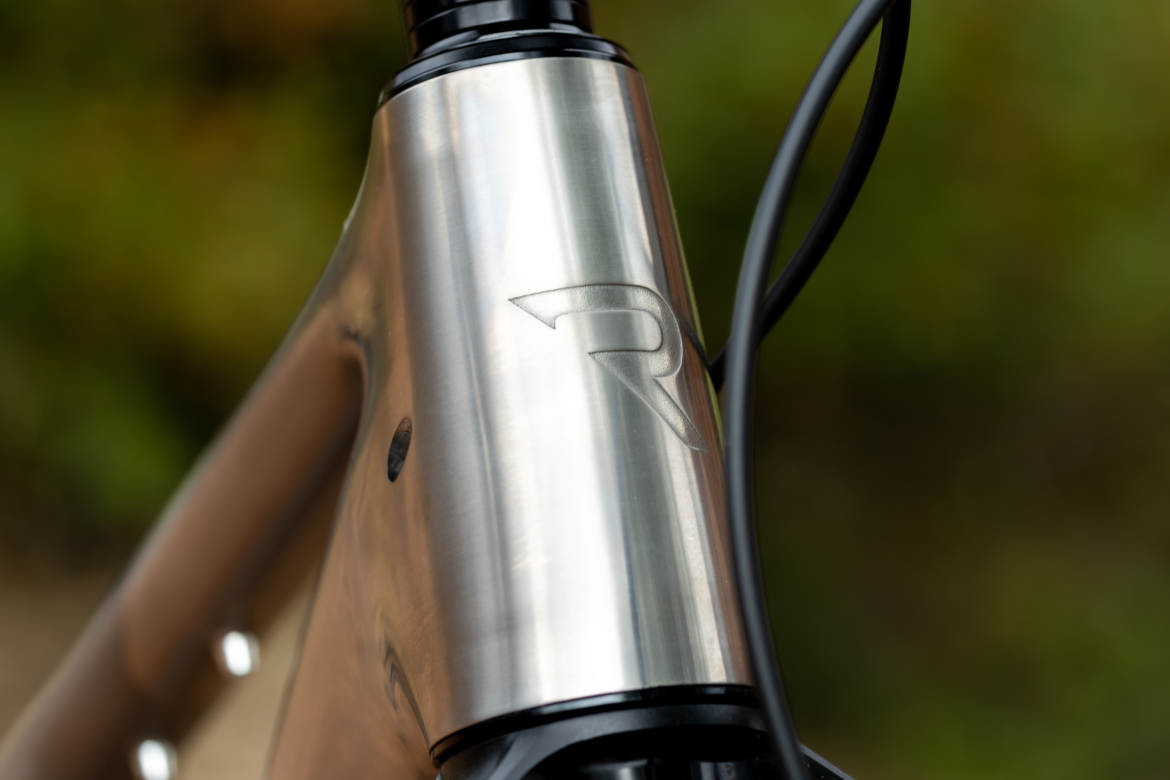 Close-up of a bicycle's metallic frame, highlighting the smooth surface and an embossed logo. The background is softly blurred, featuring natural outdoor colors.