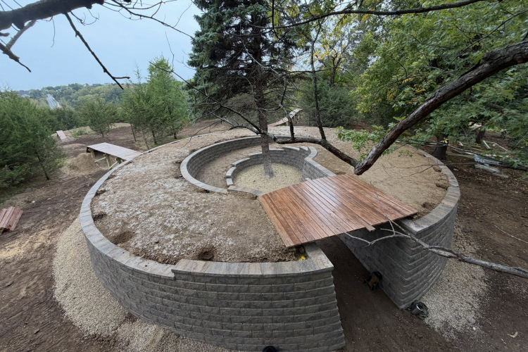 Aerial view of a circular stone structure surrounded by trees, featuring a wooden platform extending over one side and a gravel pathway leading to the entrance. The area is partially cleared with some wooden planks visible in the background, indicative of ongoing construction or landscaping.