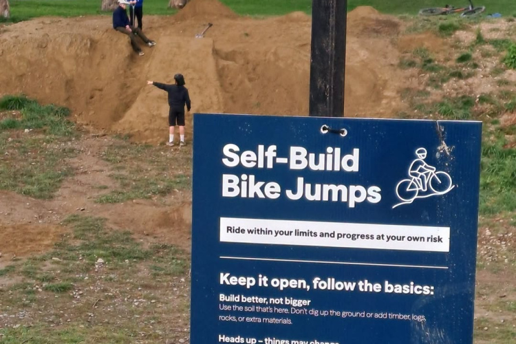 A park scene featuring a sign for "Self-Build Bike Jumps," providing safety guidelines and encouragement for riders to build and maintain bike jumps. In the background, two children are playing on a dirt ramp while a third child stands beside them. Green trees and residential houses are visible in the background, under a partly cloudy sky.
