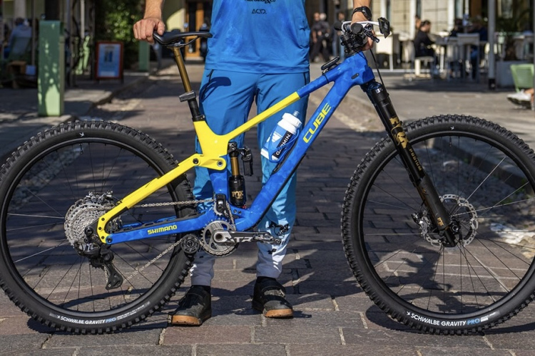 A person stands next to a vibrant blue and yellow mountain bike on a cobblestone path, with green decor and outdoor seating visible in the background. The bike features a Shimano drivetrain and Schwalbe Gravity Pro tires, showcasing its high-performance design. The rider is dressed in a blue outfit and sandals.