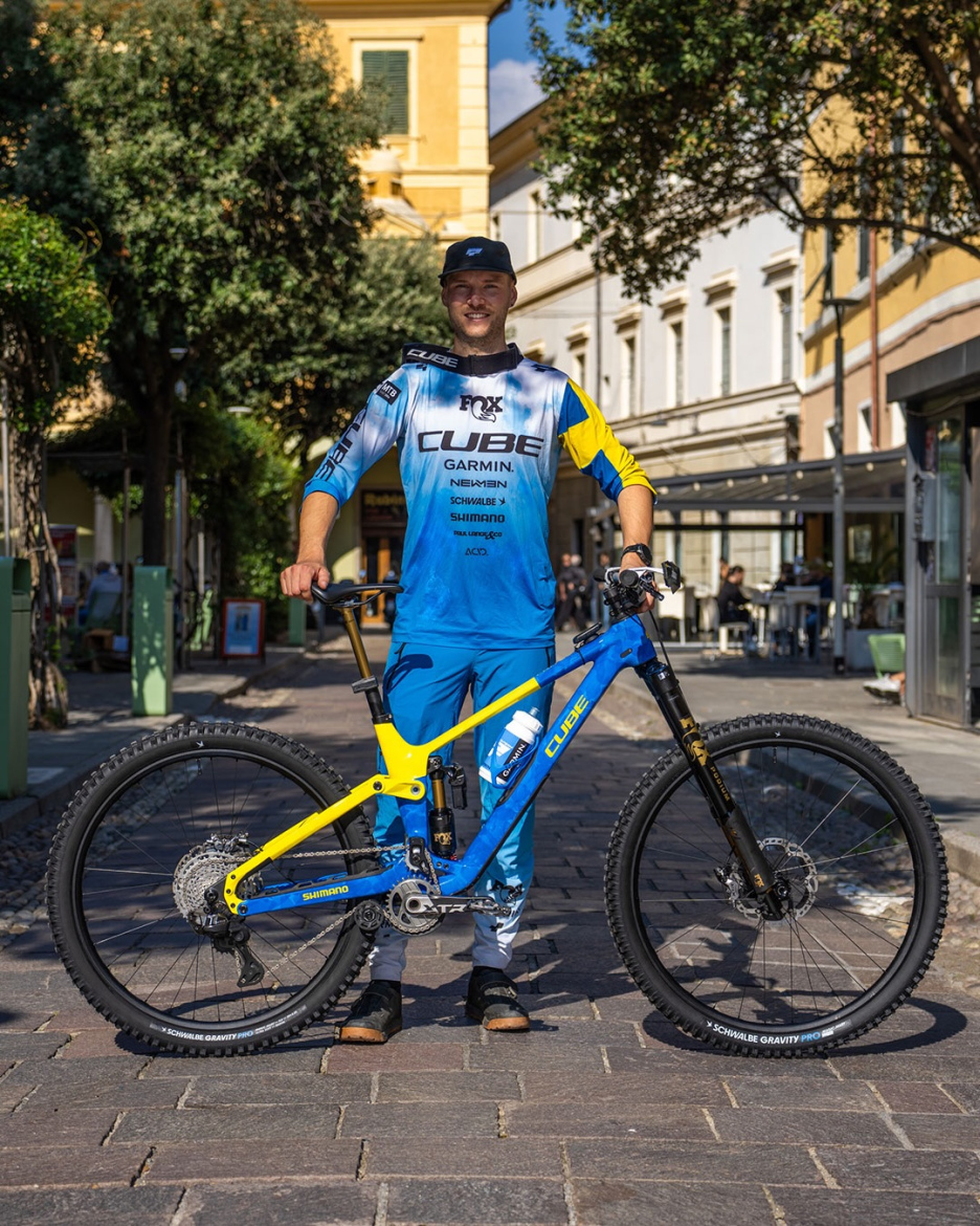 A mountain biker in a blue and yellow racing outfit stands beside a blue and yellow bicycle on a cobblestone street. The background features trees and buildings, suggesting a lively outdoor setting. The biker is smiling, wearing a black cap, and has a water bottle attached to the bike.