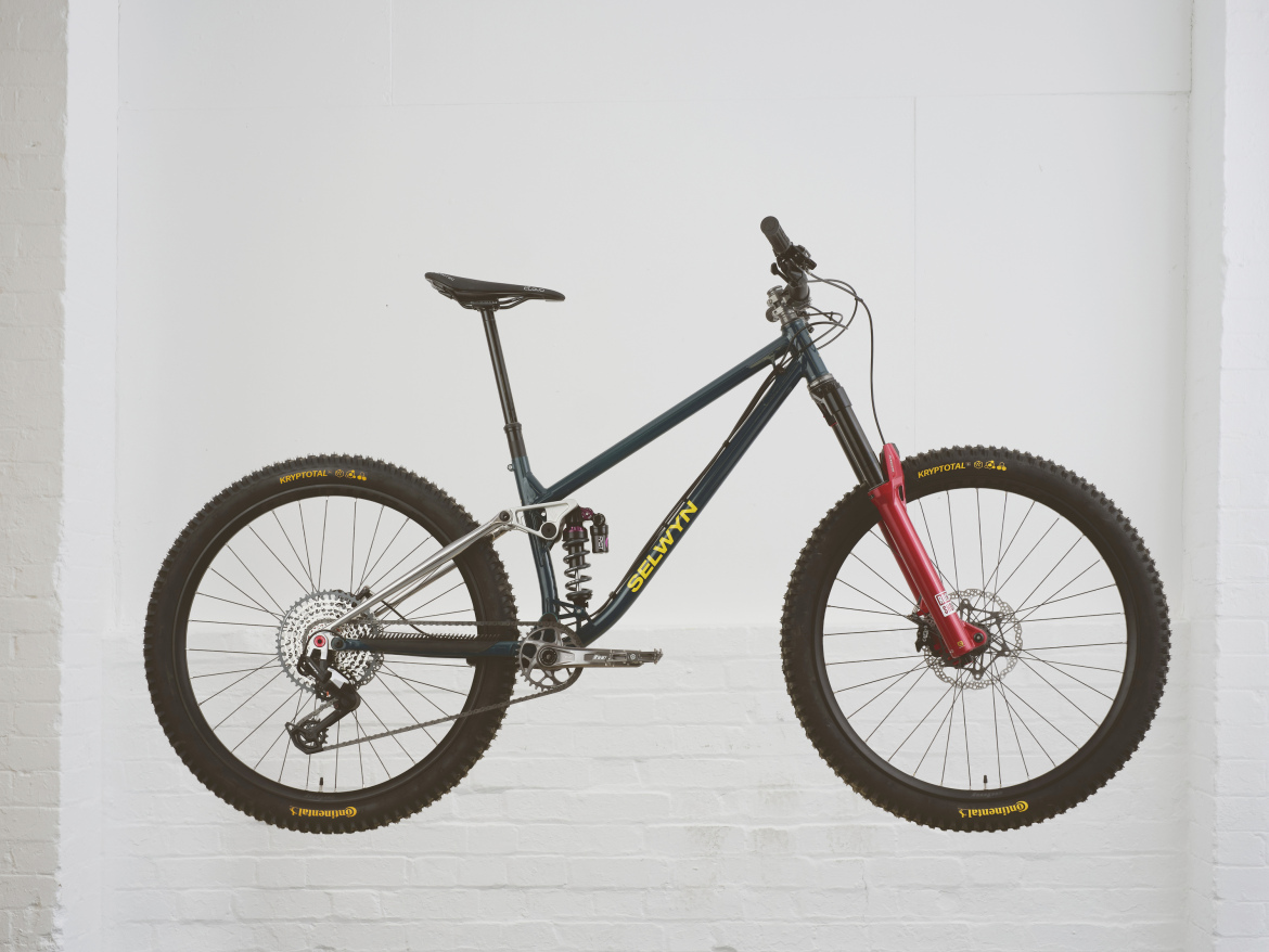 A mountain bike suspended against a white wall, showcasing a sleek design with a dark frame, red front fork, and chunky tires. The bike features a rear shock absorber and multiple gears, highlighting its suitability for off-road riding and challenging terrains.