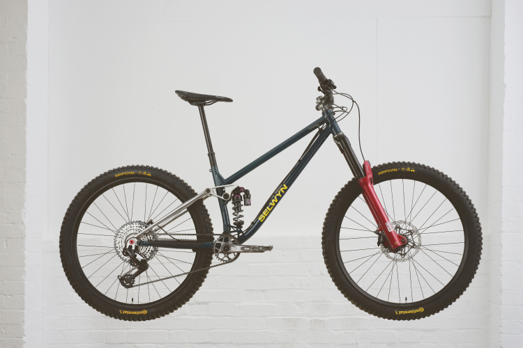 A mountain bike suspended against a white wall, showcasing a sleek design with a dark frame, red front fork, and chunky tires. The bike features a rear shock absorber and multiple gears, highlighting its suitability for off-road riding and challenging terrains.