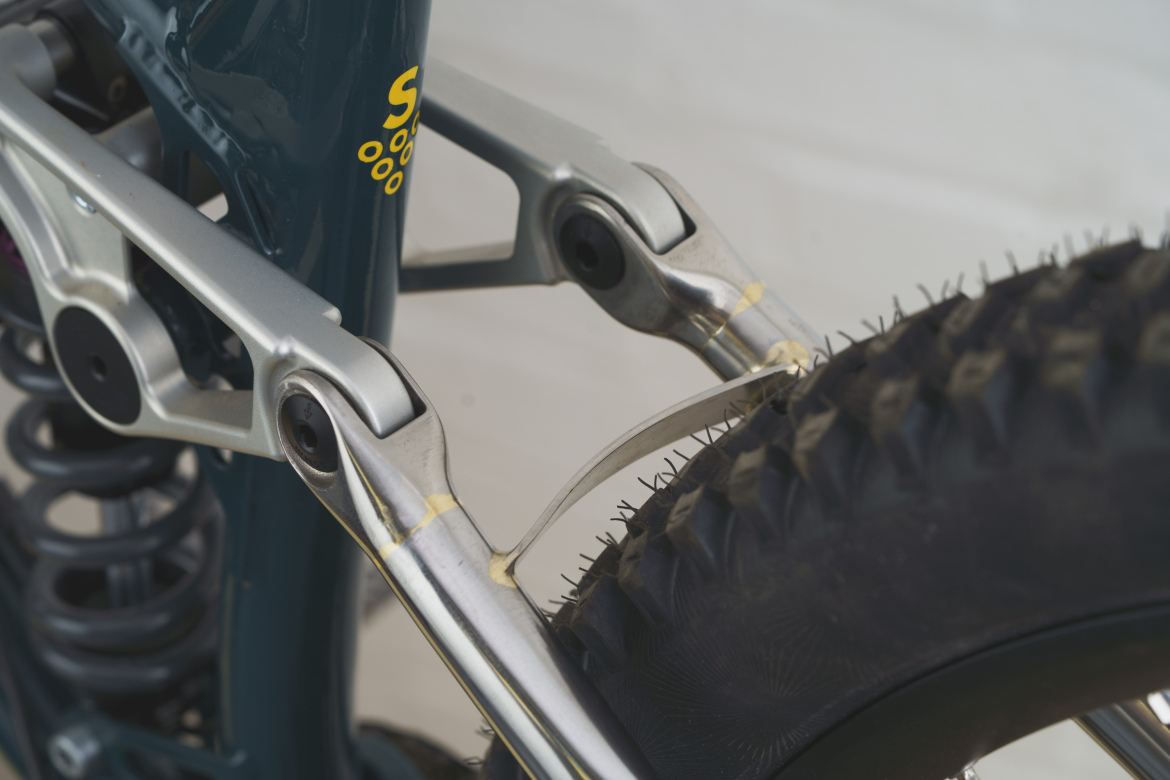 Close-up of a bicycle's rear suspension system, featuring metallic linkage components and a tire. The image highlights the intricacies of the suspension mechanism and the texture of the tire tread against a neutral background.