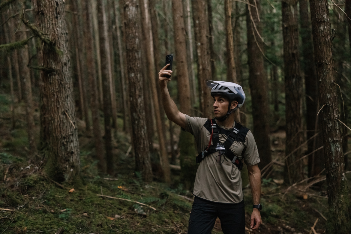 A person wearing a bike helmet and a harness takes a selfie in a dense forest filled with tall trees and greenery.