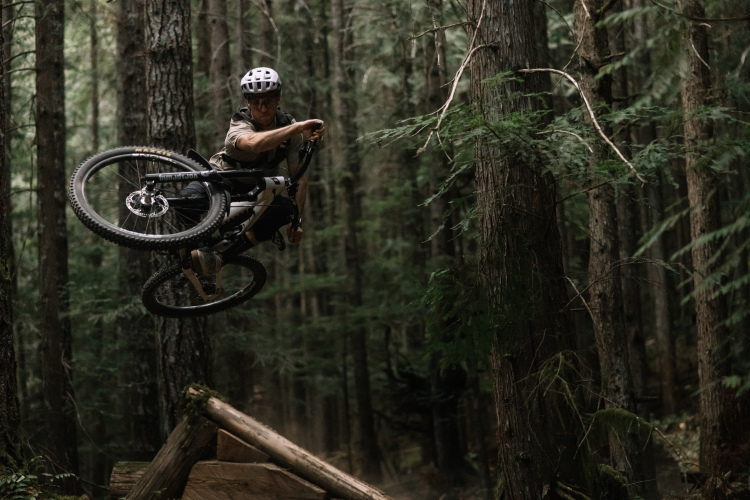 A mountain biker performs a jump over a wooden ramp in a dense forest, capturing mid-air action with the bike elevated and trees surrounding the scene.