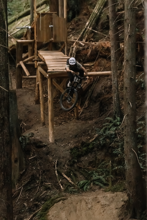 A mountain biker performing a jump off a wooden launch ramp in a forested area, surrounded by trees and natural terrain.