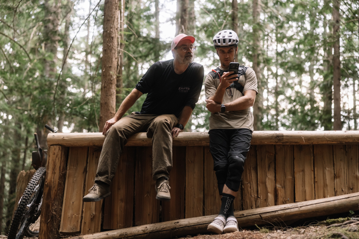 Two men are sitting on a wooden structure in a dense forest. One man, wearing a black shirt and glasses, looks over the shoulder of the other man, who is in a light-colored shirt and wearing a helmet. The second man is focused on his phone. In the background, tall trees and a mountain bike are visible.