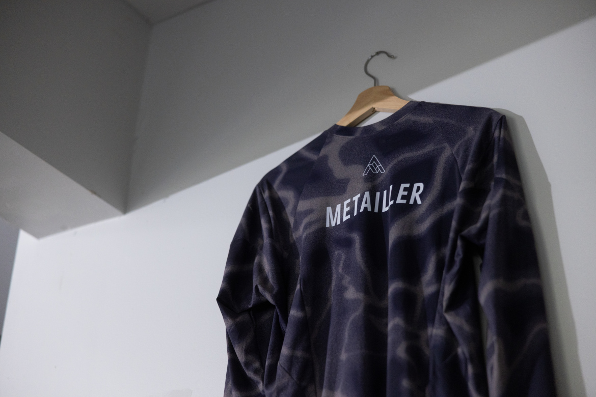 A long-sleeved shirt with a camouflage pattern hanging on a wooden hanger. The shirt features the word "METAILLER" printed in bold white letters on the back, along with a small logo above it. The background is a simple, neutral wall.