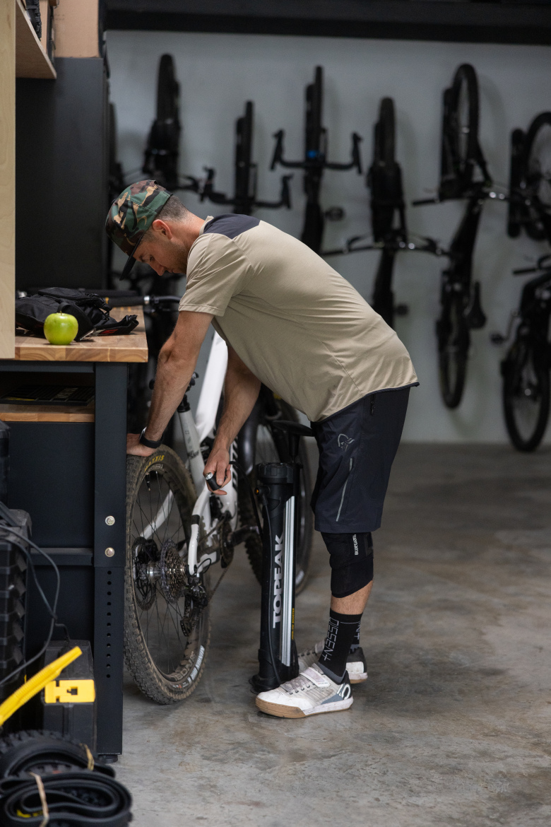 A person in casual athletic wear is working on a bicycle in a garage or workshop. They are crouched down, inspecting the bike's tire while using a hand pump. A green apple sits on a wooden workbench nearby, amidst various tools and equipment. The background features multiple bicycles mounted on a wall.