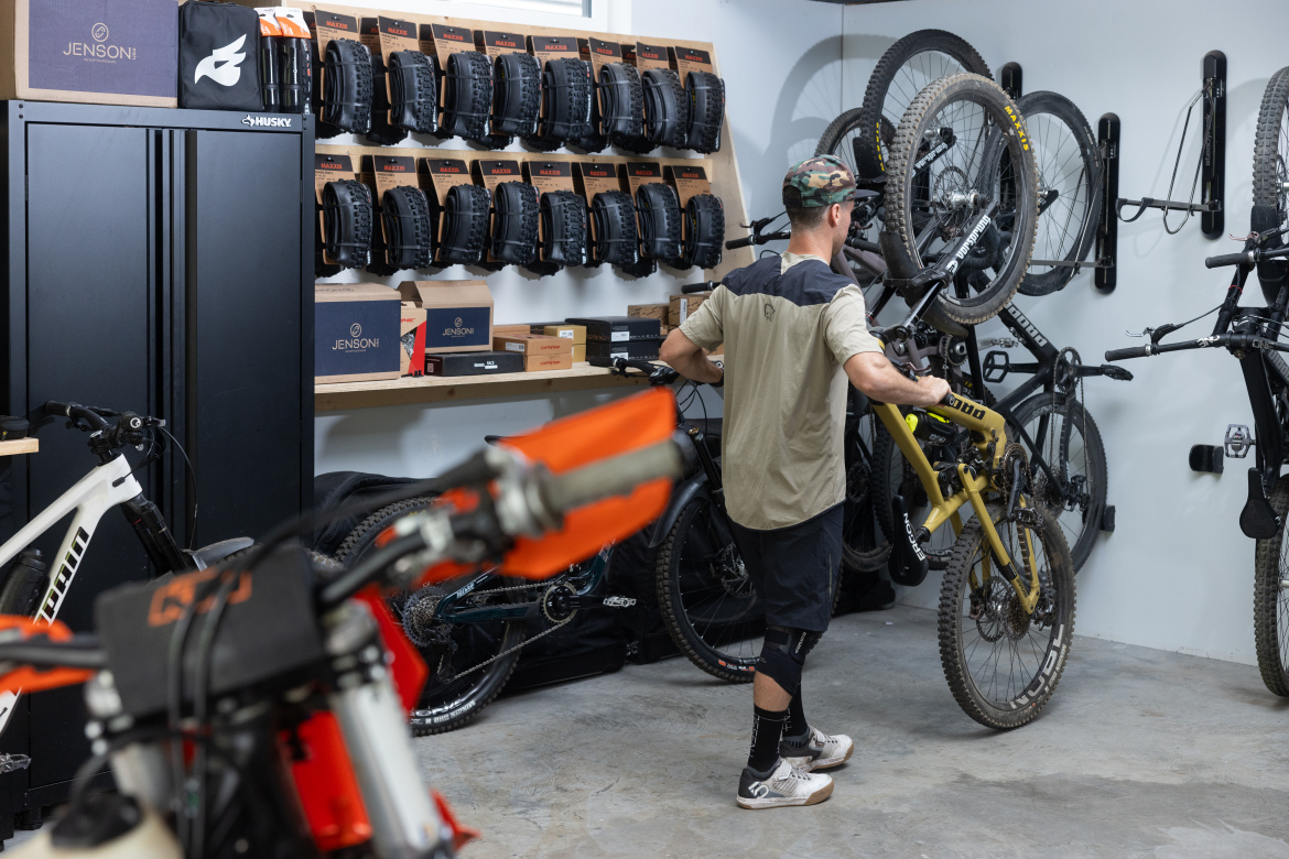 A person in a beige and black shirt is seen in a bike storage area, handling a yellow mountain bike. The walls display several mounted bicycles and a collection of bike tires. Shelves hold various bike-related items and boxes, with a black storage cabinet visible in the background. The setting has a clean, organized appearance, typical of a bike enthusiast's workshop.