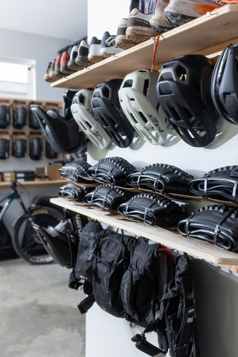 A well-organized garage space featuring a wooden shelf with various pairs of shoes on top and a selection of bicycle helmets hanging below. On another shelf, several sets of bike tires or accessories are neatly arranged. In the background, a bicycle stands against the wall, with additional gear and equipment visible, creating a functional environment for biking enthusiasts.