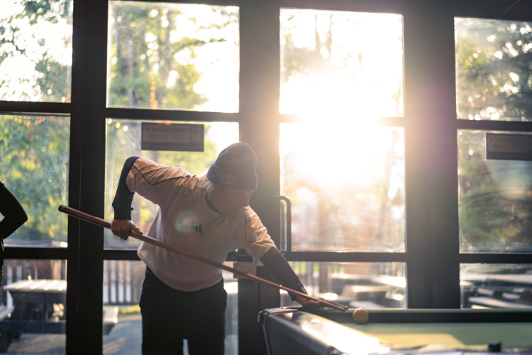 A person wearing a white shirt and a beanie is preparing to take a shot while playing pool in a warmly lit room. Sunlight filters through large windows, casting a glow on the scene, with outdoor tables visible in the background.