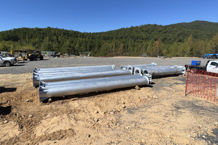 Several large, silver metal pipes stacked horizontally on a construction site with a gravel surface, surrounded by a dirt area. In the background, green hills and trees are visible under a clear blue sky. A few pieces of construction equipment and vehicles can be seen further back.