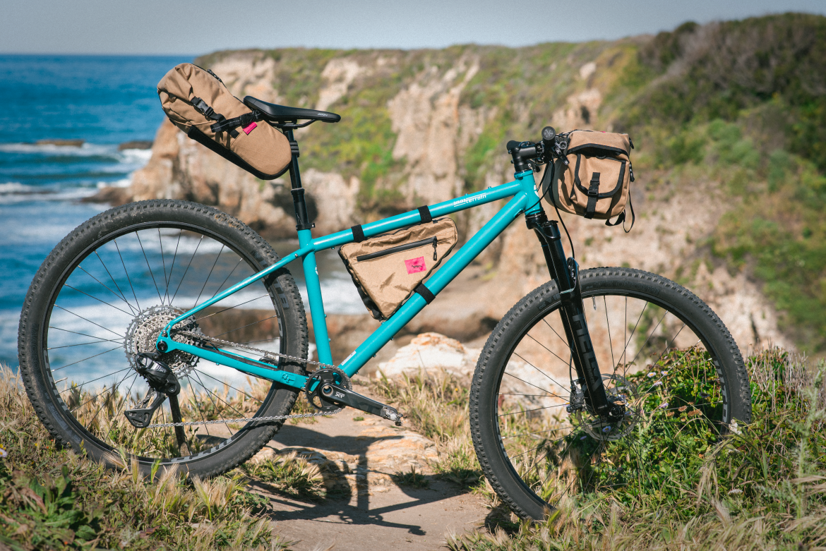 A blue mountain bike with brown accessories, including a saddle bag and frame bag, is positioned on a trail overlooking the ocean, with rocky cliffs and greenery in the background.