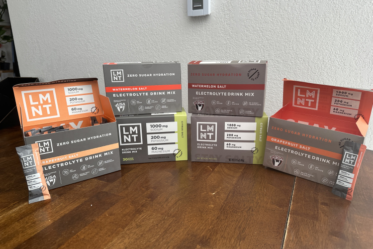 Assorted boxes of LMNT electrolyte drink mix, featuring Watermelon and Grapefruit flavors. Each box highlights zero sugar hydration with specified amounts of sodium, potassium, and magnesium. The products are displayed on a wooden table, showcasing individual stick packs.