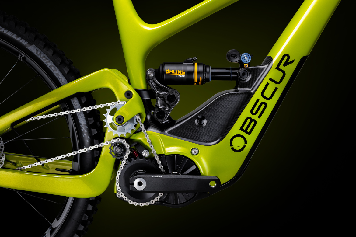 A close-up view of a vibrant green bike frame featuring a carbon fiber design. The image highlights the intricate details of the bike's suspension system, including an Öhlins shock absorber, along with the chain and gear mechanism. The background is a smooth gradient that accentuates the bike's features.