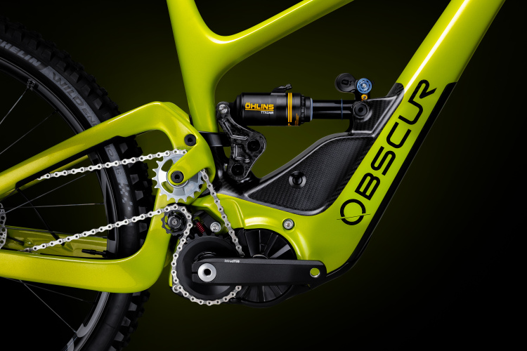 A close-up view of a vibrant green bike frame featuring a carbon fiber design. The image highlights the intricate details of the bike's suspension system, including an Öhlins shock absorber, along with the chain and gear mechanism. The background is a smooth gradient that accentuates the bike's features.