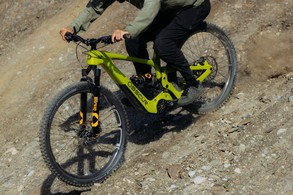A person riding a bright green mountain bike downhill on a gravel trail, kicking up dust and small rocks. The cyclist is dressed in casual outdoor clothing, with a focus on the bike's sleek design and fat tires suitable for rugged terrain.