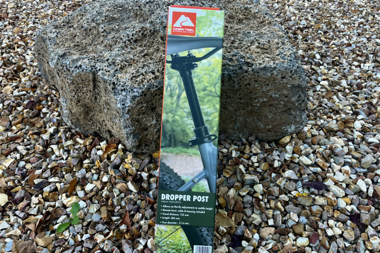 Package of a dropper post featuring features such as adjustable saddle height and travel distance, displayed on a bed of gravel with a large rock in the background.