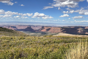 A panoramic view of a rugged landscape featuring distinctive red rock formations and mesas under a blue sky adorned with fluffy white clouds. In the foreground, green shrubs and grasses lead down to the valley below, showcasing the desert terrain and vast open spaces.