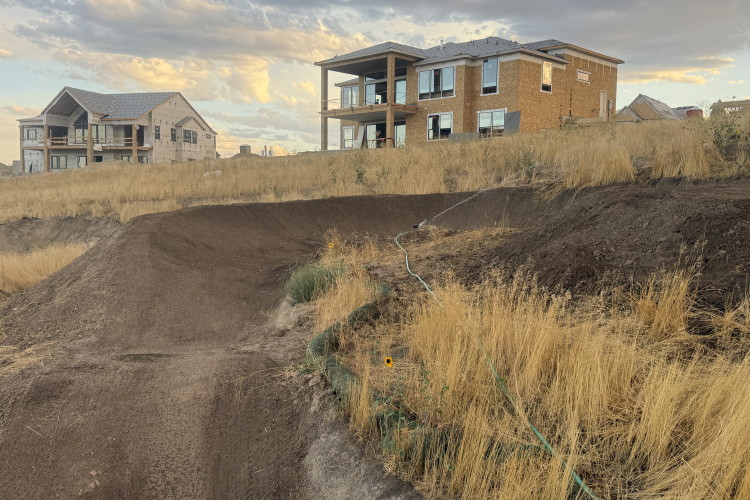 A construction site featuring two houses, one completed and one under construction, situated on a grassy hillside. A dirt ramp is visible in the foreground, with patches of dry grass and a water hose running through the area, and a cloudy sky overhead.