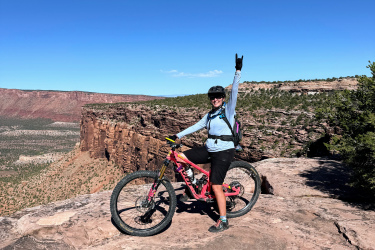A mountain biker stands on a rocky ledge with a scenic view of the landscape below, raising one hand in a victory gesture. The biker is wearing a helmet and a light blue jacket, and a pink mountain bike rests beside them. In the foreground, there is a small puddle reflecting the bike. The sky is clear and blue, highlighting the outdoor adventure setting.