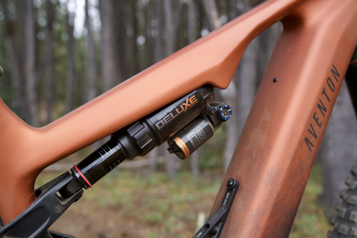 Close-up of a mountain bike frame featuring a RockShox Super Deluxe shock, showcasing the sleek copper finish with a textured pattern. The background includes blurred trees, suggesting an outdoor setting.