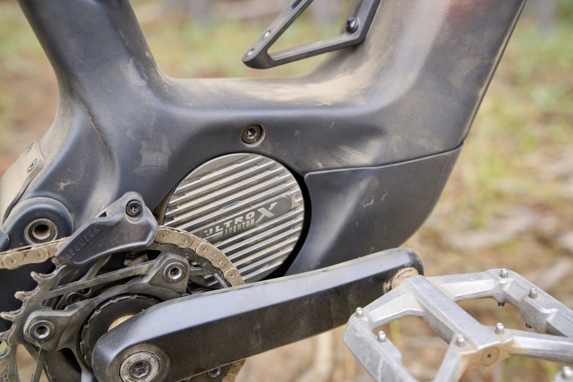 Close-up of the drivetrain and motor area of a mountain bike, showcasing a dusty black frame, chain, and a motor casing with horizontal grooves labeled "ULTRON X AVENTON." The bike appears to have been used on rugged terrain, evident from the dirt and scratches.