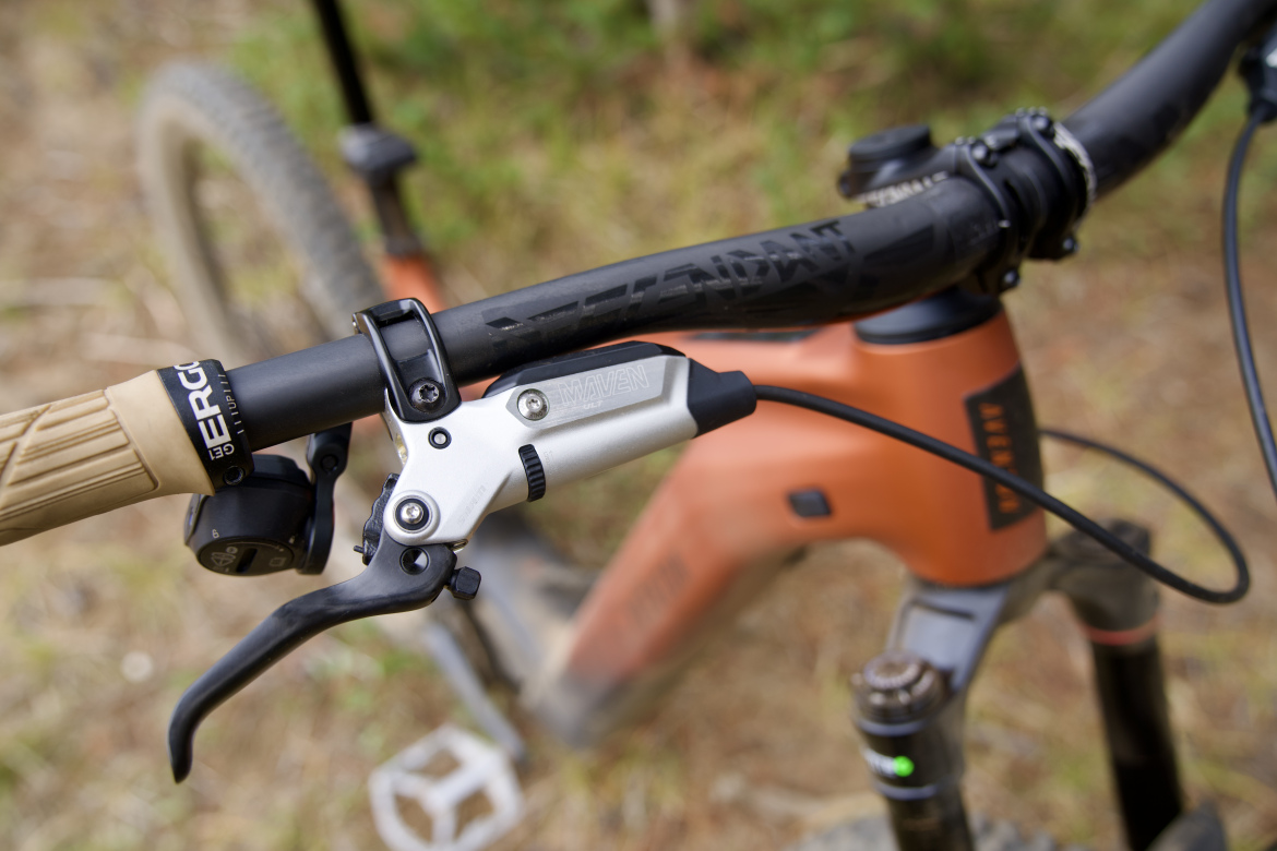 Close-up of a mountain bike handlebar featuring a silver hydraulic brake lever, beige ergonomic grips, and a gear shifter. The background shows the bike frame in shades of orange, indicating an outdoor setting.