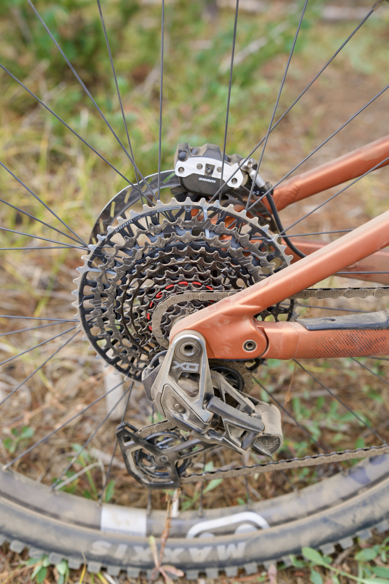 Close-up view of a bicycle's rear derailleur and cassette, showcasing a detailed arrangement of gears and chain. The bicycle features an orange frame, with visible dirt on the components, set against a natural background with grass and foliage.