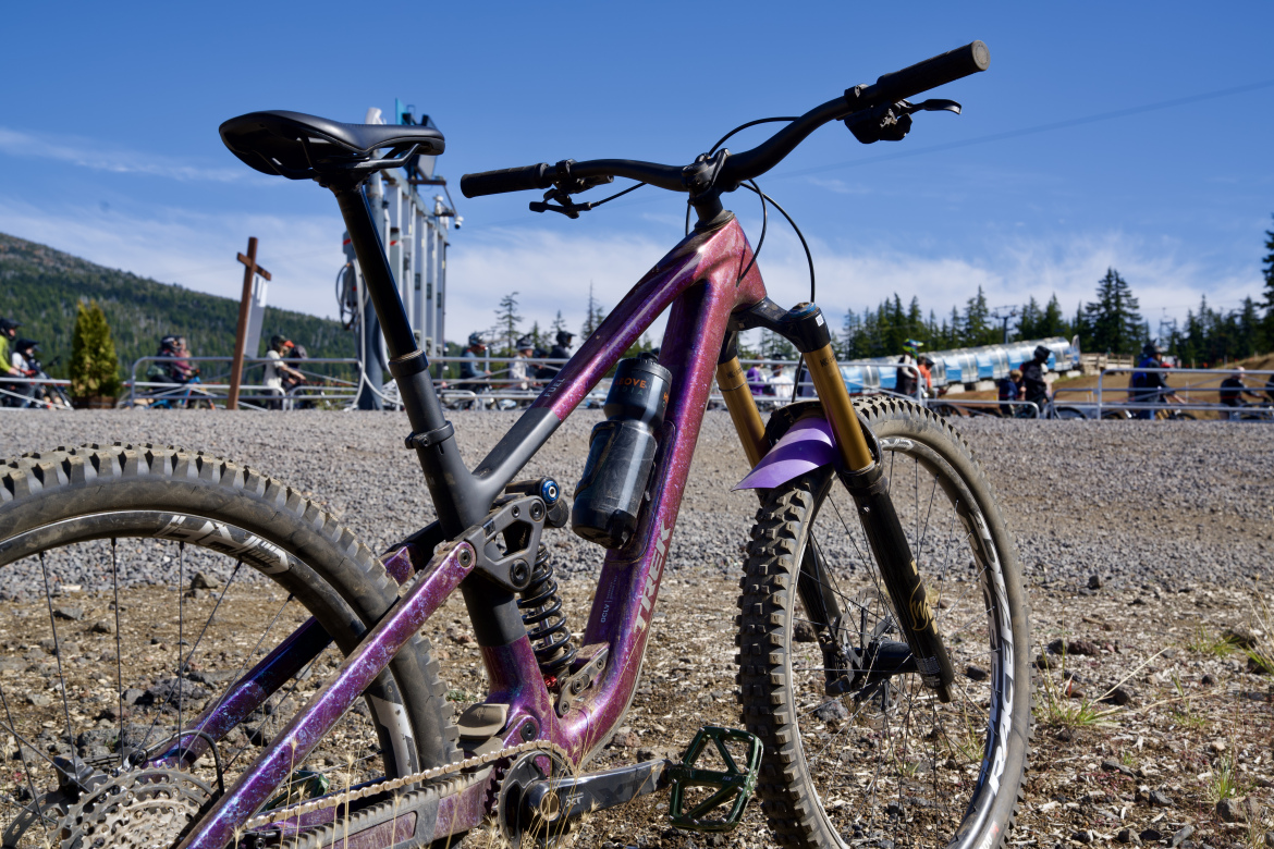 A close-up view of a colorful mountain bike with a purple and black frame, featuring a water bottle mounted on the side. The background shows a gravel area with people and a ski lift, surrounded by trees and a blue sky.
