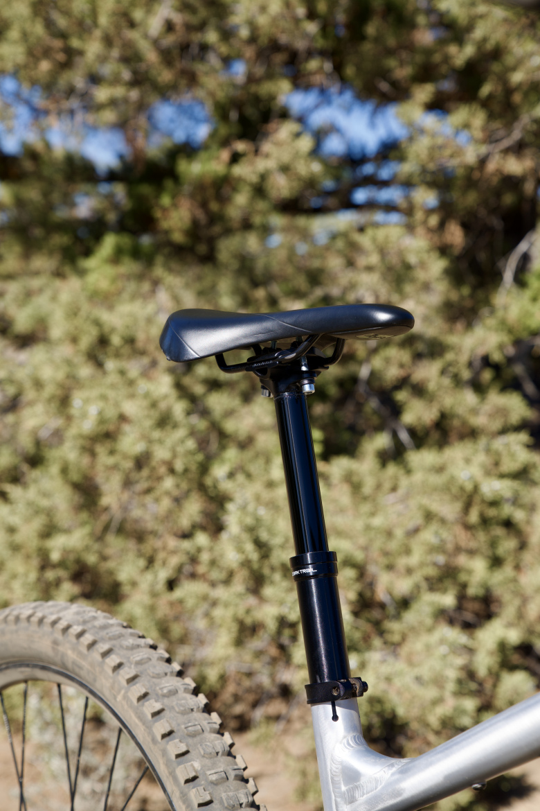 Close-up of a mountain bike seat post and saddle, set against a blurred background of greenery. The bike features a black dropper post and a textured black saddle, highlighting its design intended for off-road cycling. The bike's wheel, with a knobby tire, is partially visible in the frame.