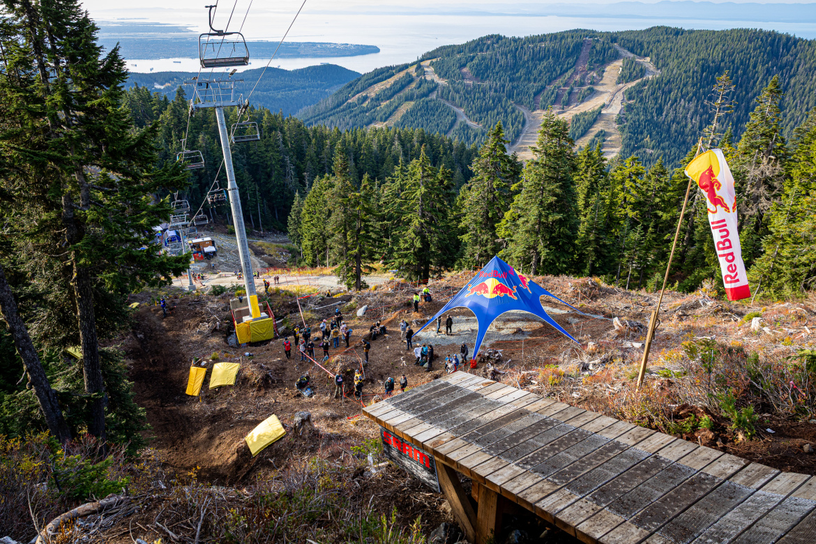 A scenic view from a mountain overlook, featuring a ski lift and a gathering area with a large Red Bull tent. In the foreground, a wooden ramp leads down the slope, while riders and spectators mill about in a rocky area surrounded by lush green trees. The backdrop showcases rolling hills and a body of water, under a clear blue sky.