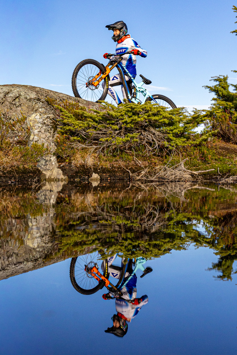 A mountain biker stands on a rock with a bicycle beside them, surrounded by greenery. The scene is reflected in calm water below, with a clear blue sky above. The biker wears a helmet and colorful cycling gear, showcasing a dynamic outdoor environment.