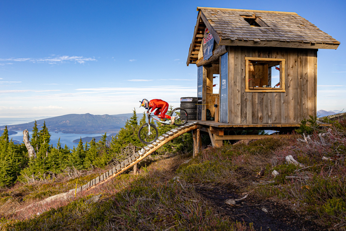 A mountain biker in a red outfit jumps off a wooden ramp leading from a small cabin set in a forested area, with a scenic view of mountains and a body of water in the background under a clear blue sky.