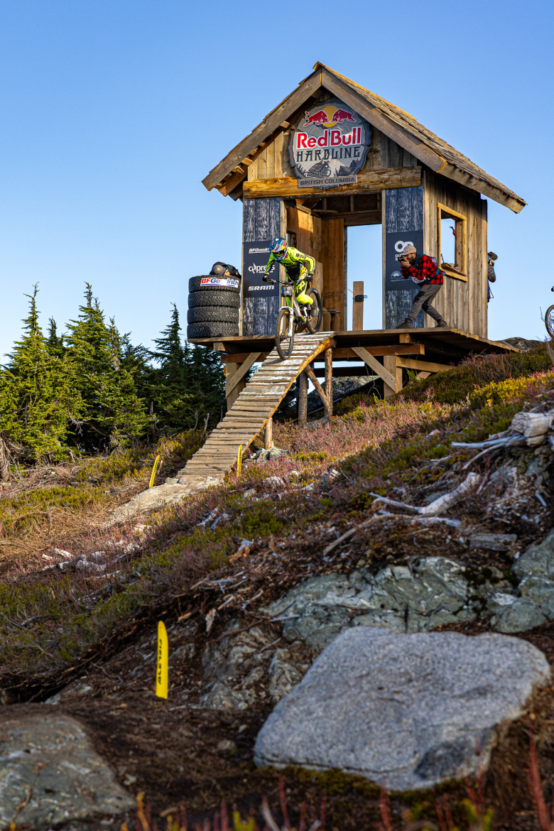 A mountain biker in bright yellow gear descends a wooden ramp from a small structure branded with the Red Bull Hardline logo. Trees surround the scene, and a spectator in a red plaid shirt watches from the platform. The sun is shining, creating a vibrant outdoor atmosphere, with rocky and hilly terrain in the foreground.